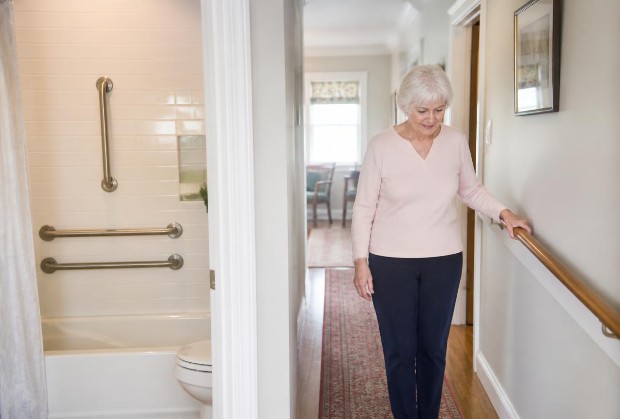 Older woman using a hallway handrail at home, with bathroom grab bars installed nearby
